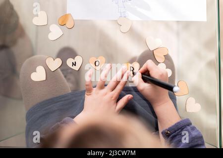 Vista dall'alto di raccolto anonimo capretto scrittura iscrizione su piccoli cuori mentre si siede al tavolo di vetro in camera Foto Stock