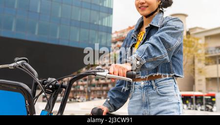 Corto irriconoscibile colombiana giovane donna indossando abbigliamento alla moda denim e casco protettivo in piedi con bicicletta vicino moderno edificio di vetro durante AC Foto Stock