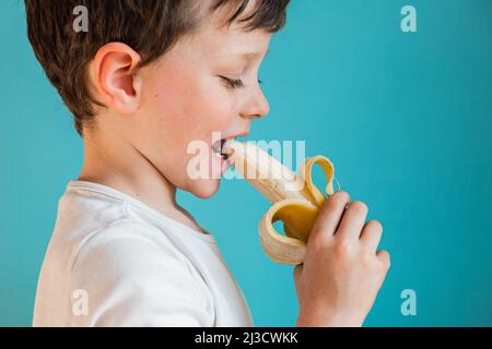 Vista laterale del ragazzo felice mangiare giallo maturo pelato gustoso banana con sorriso mentre in piedi su sfondo blu in studio chiaro Foto Stock