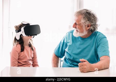 Ragazza carina in moderni occhiali VR esplorare il cyberspazio mentre si siede vicino a nonno barbuto nella stanza luminosa di casa Foto Stock