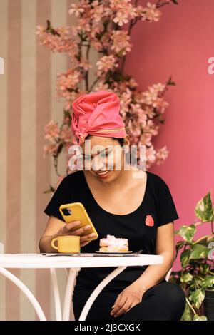 Felice ispanica femmina in foulard seduta al tavolo e navigare social media sul cellulare il giorno del fine settimana in caffetteria Foto Stock