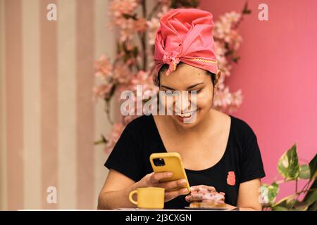 Felice ispanica femmina in foulard seduta al tavolo e navigare social media sul cellulare il giorno del fine settimana in caffetteria Foto Stock