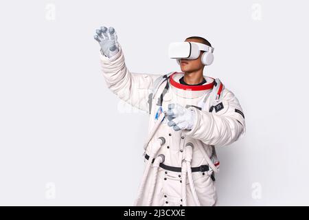 Cosmonaut maschile colombiano in uniforme interagendo con il cosmo virtuale in occhiali e raggiungendo mano in avanti in studio su sfondo bianco Foto Stock