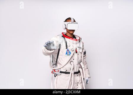 Cosmonaut maschile colombiano in uniforme interagendo con il cosmo virtuale in occhiali e raggiungendo mano in avanti in studio su sfondo bianco Foto Stock