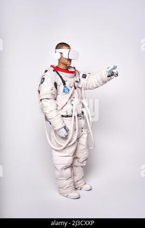 Cosmonaut maschile colombiano in uniforme interagendo con il cosmo virtuale in occhiali e raggiungendo mano in avanti in studio su sfondo bianco Foto Stock