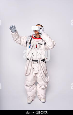 Cosmonaut maschile colombiano in uniforme interagendo con il cosmo virtuale in occhiali e raggiungendo mano in avanti in studio su sfondo bianco Foto Stock