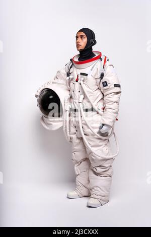 Corpo pieno di astronauta maschio colombiano in spacesuit con casco in mano in piedi in studio su sfondo bianco e guardando via Foto Stock