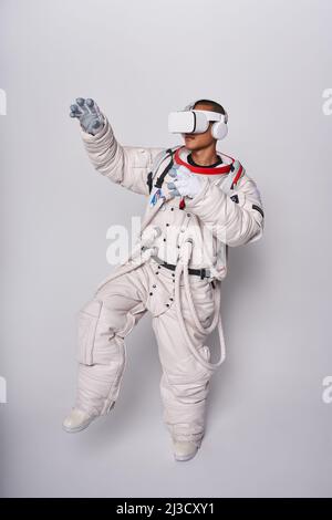 Cosmonaut maschile colombiano in uniforme interagendo con il cosmo virtuale in occhiali e raggiungendo mano in avanti in studio su sfondo bianco Foto Stock