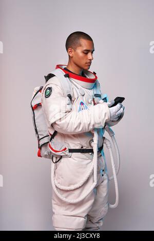 Fiducioso colombiano giovane maschio cosmonaut indossando spacesuit navigazione cellulare in studio su sfondo bianco Foto Stock