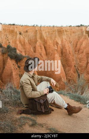 Vista laterale del giovane uomo in abiti casual e cappello con macchina fotografica seduta su terreno asciutto e ammirare il crinale di montagna durante il viaggio in campagna Foto Stock