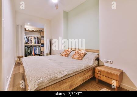 Comodo letto con cuscini posto vicino armadietto in legno in camera da letto spaziosa e luminosa con armadio aperto con vari vestiti a casa Foto Stock