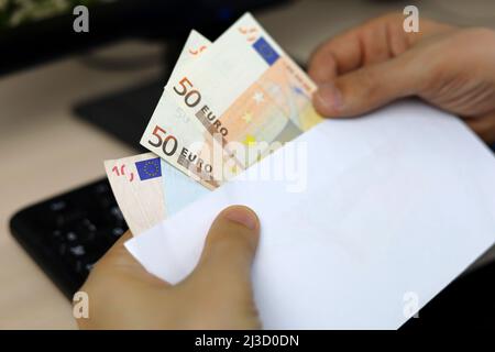 Busta con banconote in euro in mani maschili. L'uomo estrae denaro da una busta sullo sfondo della tastiera del PC, salari, bonus o concetto di tangente Foto Stock