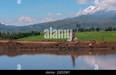 Bella donna ispanica cammina da sola con un abito fiorito e cappello lungo una strada sterrata che si riflette in una laguna con il vulcano Cayambe sullo sfondo Foto Stock