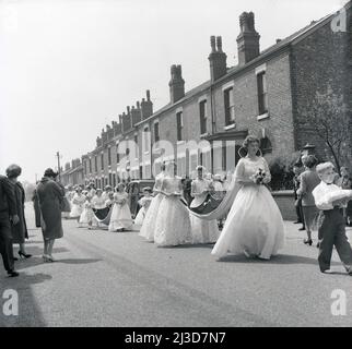 1961, storica, processione del giorno di maggio, due ragazze giovani in abiti lunghi tengono il treno pesante del tessuto della città nuova incoronata regina di maggio, mentre camminano lungo una strada di case a schiera vittoriane a Stockport, Manchester, Inghilterra, Regno Unito. Foto Stock