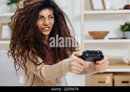 Concentrato overjoyed abbastanza abbronzato curly latino lady giocare in gioco con joystick gamepad sedersi su sedia in casa interni moderni guardare da parte. Spazio di copia Foto Stock