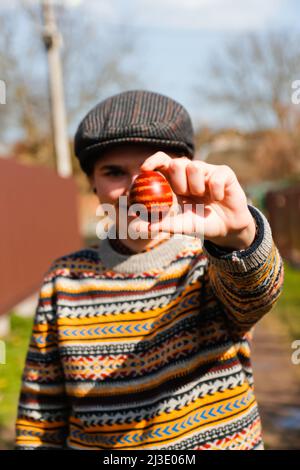 Defocus giovane donna ucraina in cappello che tiene un uovo rosso colorato su sfondo naturale. Pasqua, Ucraina. Uova dipinte di mestiere. Pysanka o krashanka. Fuori Foto Stock