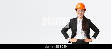 Professionista sorridente ingegnere asiatico di costruzione, architetto in tuta nera e casco di sicurezza guardando felice al lavoro, in piedi soddisfatti Foto Stock