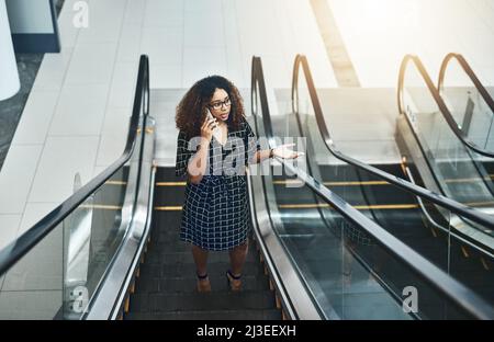 Realizzare piani aziendali in movimento. Colpo ad angolo alto di una giovane donna d'affari attraente che prende una telefonata mentre salendo su una scala mobile in un moderno Foto Stock