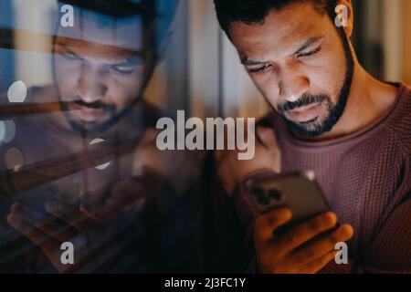 Il giovane uomo con uno smartphone sembra triste, in piedi dalla finestra di sera a casa. Foto Stock