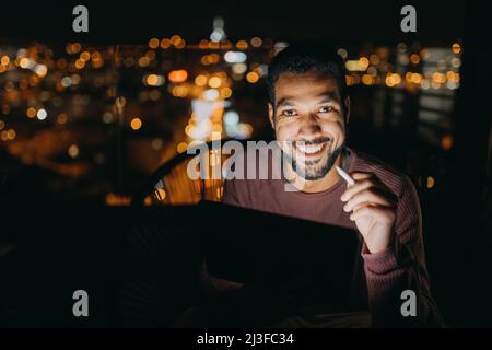 Giovane afroamericano seduto sul balcone con vista sulla città e con un tablet di notte Foto Stock