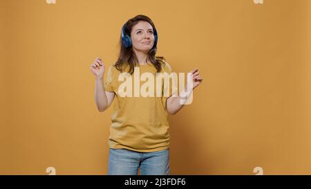 Ritratto di donna positiva pieno di energia con cuffie wireless che hanno partito e ballare da solo in studio. Donna casual che ascolta musica ottimistica sulle cuffie e fa mosse di danza stravaganti. Foto Stock