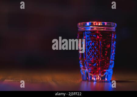 Tintura di colore rosso in vetro cristallo si erge su un tavolo di legno con illuminazione blu Foto Stock