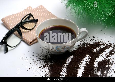 studio fotografico di caffè nero in una tazza con fondi di caffè grossolani e un occhiale marrone su sfondo bianco e verde pianta Foto Stock