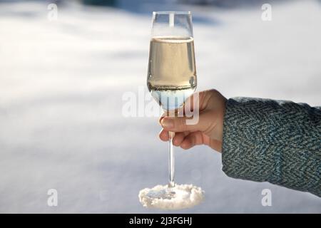 La mano di una donna in una giacca calda contiene un bicchiere di champagne coperto di neve. Vacanze invernali Foto Stock