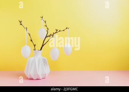 Buona vacanza di Pasqua, in arrivo primavera concetto. Rami di albero, rametti di fiori in vaso bianco o vaso con uova appese. Sfondo giallo e rosa della tabella con spazio di copia. Preparazione di Pasqua, biglietto di auguri Foto Stock