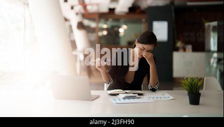 Donna asiatica molto stressata di affari seduta davanti al suo calcolatore che guarda un mucchio grande di lavoro di ufficio, mentre tiene una mano alla sua fronte Foto Stock
