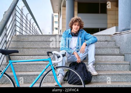 Corpo pieno di positivo maschio capelli ricci in abiti casual con cuffie sul collo seduta su scala vicino alla bicicletta per le strade della città Foto Stock