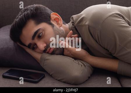 Ritratto di un giovane uomo sdraiato sul divano e guardando lo smartphone tenuto accanto Foto Stock