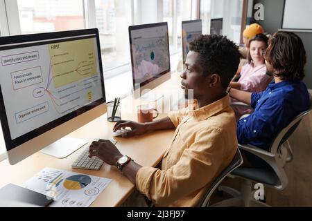 Concentrato il giovane manager afro-americano seduto a tavola nello spazio di coworking e disegno di strategia sul computer Foto Stock