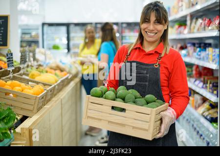Felice etnia femmina venditore con scatola di avocado in supermercato Foto Stock