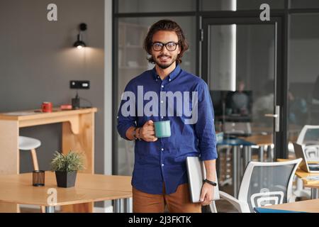 Ritratto di un giovane e sorridente sviluppatore di app per gare miste che tiene un laptop e beve caffè nello spazio di lavoro Foto Stock