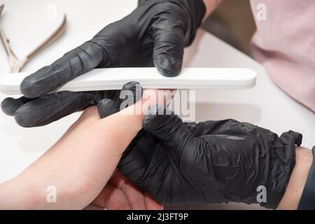 Mani gloved di un manicurist esperto che archivano le unghie di una donna giovane con una lima del chiodo. Mani durante una sessione di cura manicure in un salone di spa. Manicurista fi Foto Stock