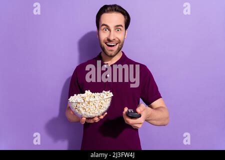 Foto di impressi uomo brunetta millennial mangiare pop corn guardare la tv indossare viola t-shirt isolato su sfondo di colore viola Foto Stock