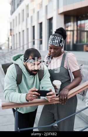 Giovani amici multietnici che si appoggiano sulla ringhiera in città e guardano i video sullo smartphone Foto Stock