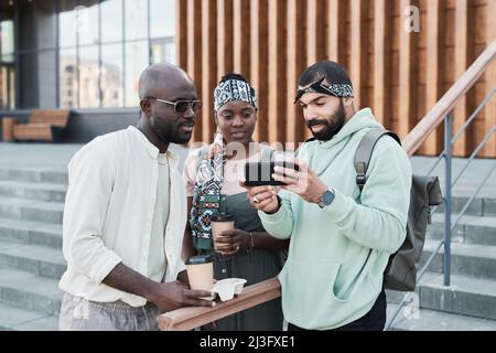 Uomo portico medio-orientale con borsa a mano che mostra foto al telefono agli amici con caffè da asporto Foto Stock