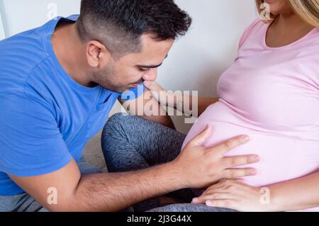 primo piano giovane uomo caucasico che parla con il bambino all'interno del ventre della moglie incinta, seduto sul letto godendo un momento di amore e di convivenza Foto Stock