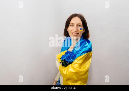 Una giovane ragazza indossa la bandiera nazionale dell'Ucraina e tiene i fiori su sfondo bianco. La ragazza ha una bandiera blu e gialla sulla guancia. Giornata dell'indipendenza dell'Ucraina. Spazio copia. Foto Stock