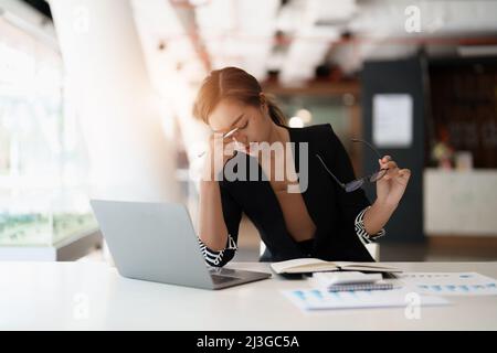 Donna asiatica molto stressata di affari seduta davanti al suo calcolatore che guarda un mucchio grande di lavoro di ufficio, mentre tiene una mano alla sua fronte Foto Stock