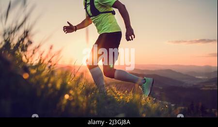Foto ritagliata di un corridore di montagna di media età vestito t-shirt brillante con uno zaino resistenza che corre in salita da pittoresche colline al tramonto Foto Stock