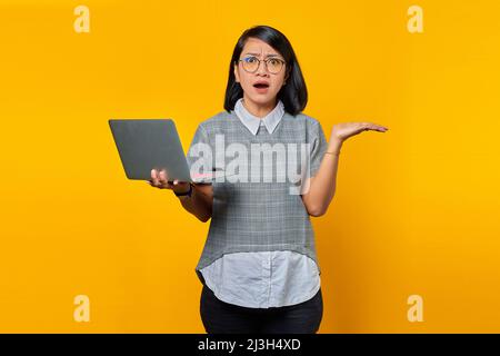 Ritratto di donna asiatica indossando occhiali in presa a sorpresa laptop con confuso volto su sfondo giallo Foto Stock