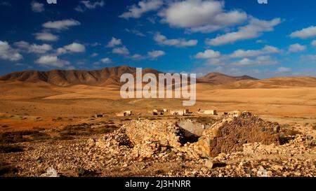 Spagna, Isole Canarie, Fuerteventura, interno dell'isola, paesaggio arido, dry. deserto-come, catena montuosa arida, cielo blu con nuvole bianche, edificio in rovina in primo piano, isolati edifici abbandonati nel terreno centrale Foto Stock