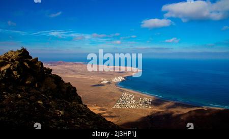 Isole Canarie, Lanzarote, isola vulcanica, costa nord-occidentale, eremo, chiesa, Ermita de las Nieves, cielo blu, grandangolo colpo, vista dall'Ermita fino alla costa occidentale, nelle rocce in primo piano, sotto si può vedere un insediamento turistico bianco, cielo blu con nuvole bianche, mare blu e verde Foto Stock