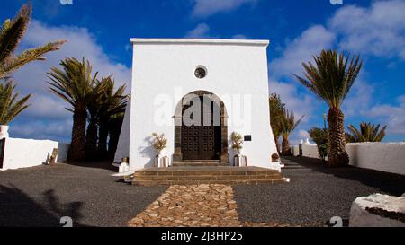 Isole Canarie, Lanzarote, isola vulcanica, costa nord-occidentale, eremo, chiesa, Ermita de las Nieves, blu cielo, edificio bianco dell'Ermita, porta grande marrone, pietra incorniciata, palme a sinistra e a destra, blu cielo con nuvole bianche Foto Stock