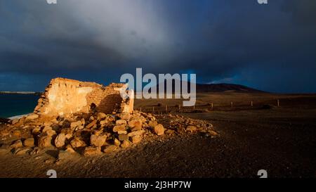 Isole Canarie, Lanzarote, isola vulcanica, atmosfera post-temporale, spiagge di Papagallo, deserta, blu cielo con nuvole grigio-bianco, luce del mattino, rovine di un vecchio edificio, pareti, sullo sfondo nero colline, cielo scuro e minaccioso, solo l'edificio è illuminato dal sole del mattino Foto Stock