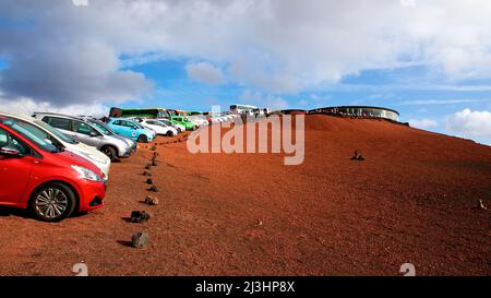 Isole Canarie, Lanzarote, isola vulcanica, Parco Nazionale di Timanfaya, paesaggi vulcanici, Beuscherzentrum, ristorante rotondo di roccia vulcanica sulla destra sullo sfondo, cielo blu sopra con nuvole bianche-grigie, di fronte ad esso auto parcheggiate in una lunga fila su terra di lava rossa Foto Stock