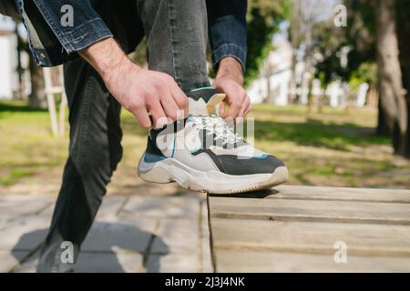 Scarpe sportive, il giovane uomo ha messo il piede sulla panca del parco e legando le scarpe, un prodotto adatto per uso quotidiano con dettagli grigi e blu Foto Stock